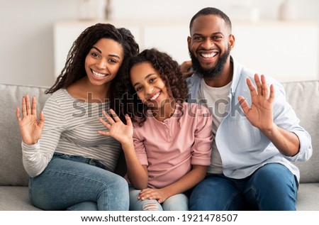 Happy Loving Family. Portrait of cheerful African American man, woman and girl sitting on the couch at home, posing for photo and waving hands to camera. Smiling young black people
