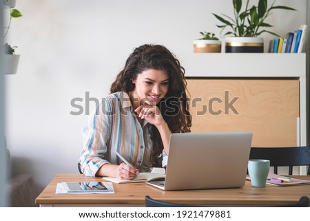 Smiling business woman teleconferencing from home. Happy businesswoman sitting and talking to colleagues in a meeting using her laptop computer and writing down notes Royalty-Free Stock Photo #1921479881