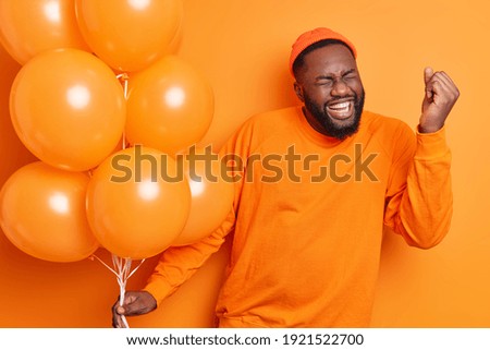 Overjoyed male student celebrates success makes yes gesture being on graduation party holds inflated balloons dressed in casual jumper isolated over orange background. People happiness concept Royalty-Free Stock Photo #1921522700