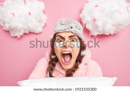 Close up shot of emotional brunette European woman yells loudly keeps mouth widely opened dressed in pajama holds pillow being angry with someone who interrupted her sleep surrounded with feathers