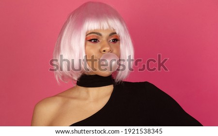 Photo of good looking young African American female with vivid make up inflates a bubble of gum  on a pink background.