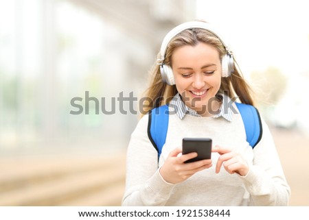 Happy student wearing headphones listening to music on smart phone walking in the street