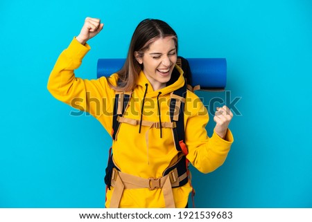 Young mountaineer woman with a big backpack isolated on blue background celebrating a victory