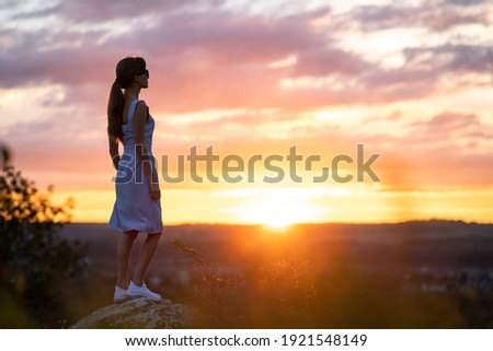 A young woman in summer dress standing outdoors enjoying view of bright yellow sunset.