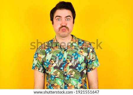 young handsome Caucasian man wearing Hawaiian shirt against yellow wall puffing cheeks with funny face. Mouth inflated with air, crazy expression.