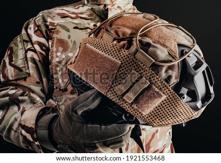 Photo of soldier in camouflaged uniform and tactical gloves holding military helmet with cover on black background.