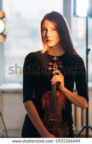 Violinist woman. Professional artist. Studio shooting. Beautiful elegant lady having photo session with violin in hands looking at camera in reflectors warm lights on background.
