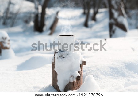 An enamelled mug with a hot drink standing on a snow-covered stump in a winter forest. Steam rises from the metal mug. Sunny day, camping concept.