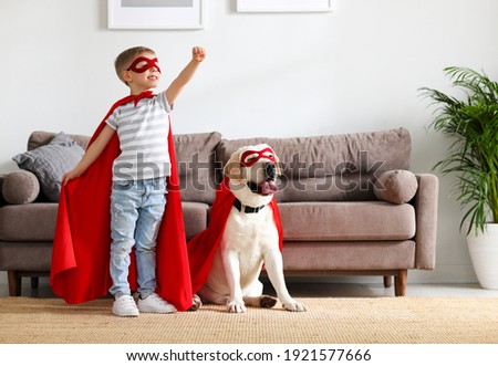 Full body of little boy in red superhero cloak and mask raising hand while playing with funny dog dressed in similar costume in living room at home Royalty-Free Stock Photo #1921577666