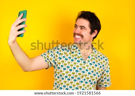 Portrait of a young handsome Caucasian man wearing Hawaiian shirt against yellow background  taking a selfie to send it to friends and followers or post it on his social media.