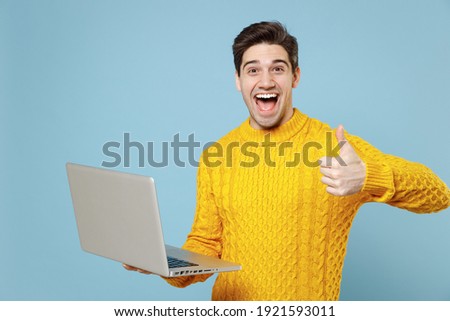 Young caucasian successful shocked surprised man 20s in casual knitted yellow sweater hold in hand using laptop pc computer show thumb up gesture like isolated on blue color background studio portrait