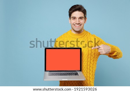 Young smiling man 20s in casual knitted yellow sweater hold in hand using point index finger on laptop pc computer with blank screen workspace area isolated on blue color background studio portrait
