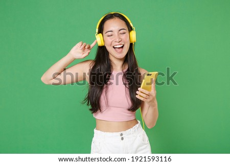 Cheerful young asian woman girl in pink clothes isolated on green background studio. People lifestyle concept. Mock up copy space. Listening music with headphones, mobile phone, sing song dancing Royalty-Free Stock Photo #1921593116