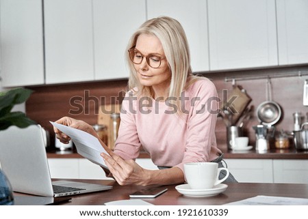 Middle aged older woman housewife reading paper letter or bill sitting in kitchen at home office, checking financial taxes fees, reviewing bank account loan rates information, medical insurance cost.