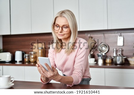 Middle aged 50 years old woman using apps ordering buying food on smartphone sitting in kitchen at home. Mature older lady holding mobile phone texting messages, browsing online services.