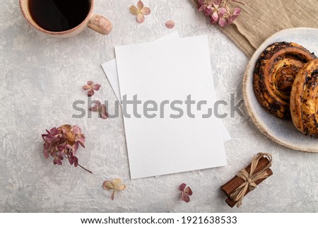 White paper invitation card, mockup with hydrangea flowers, bun, cup of coffee, linen textile, cinnamon on gray concrete background. Blank, flat lay, top view, still life, copy space.