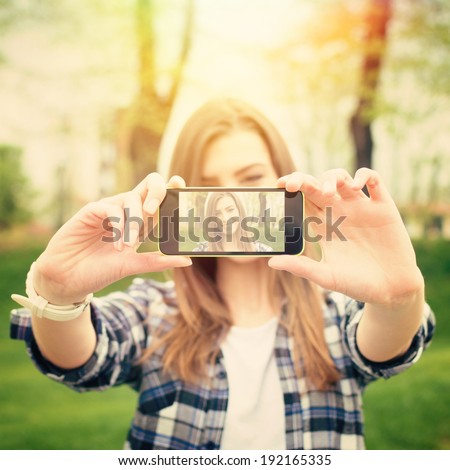 Closeup of beautiful young woman taking a selfie photo with smart phone outdoors on sunny summer day. Instant filter. Square image.
