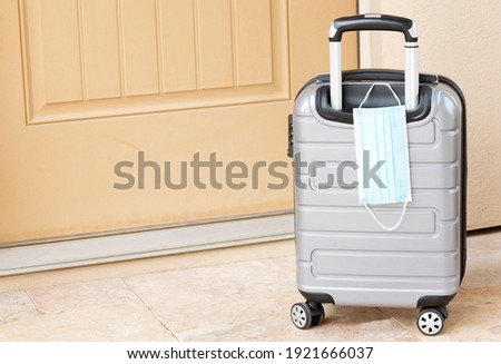 Pandemic travel seen in gray suitcase, and protective face mask by tan door and ready for departure