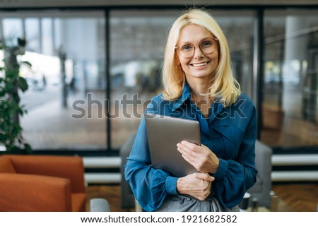 Joyful blonde-hair business woman in eyeglasses is looking at the camera, holding laptop in arms and smiling. Beautiful middle aged lady in stylish wear stands in modern office