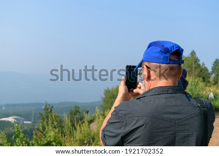 An older man standing on a mountain takes a photo on a smartphone. View from the back, close-up. Green grass and foliage in summer, sky.