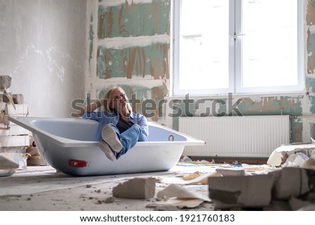 Renovation apartment. Creative story young happy woman sits in bathtub in the middle of the room. Empty walls, repairs house with their own hands. Royalty-Free Stock Photo #1921760183