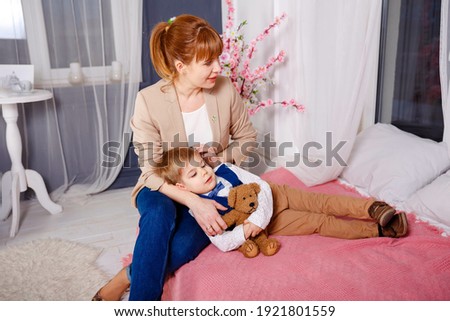 Young mother holding her little sick boy. Sick child with high fever laying in bed. Mommy checking on sick son laying in bed. Sleeping boy laying in lap of mom. Mothercare for a sick child with fever.