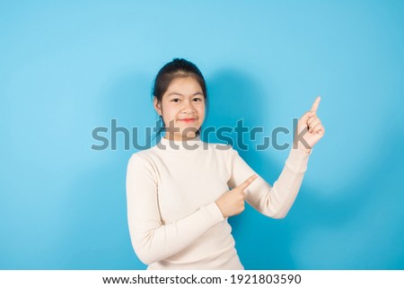 Portrait of smiling Asian teenager  joyfully and pointing finger to the side, Wears in white knitted sweater isolated over blue background with copy space