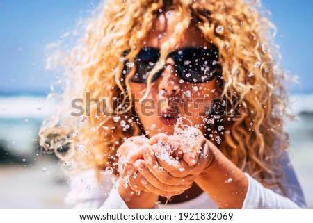 Close up of woman blowing sea water from hands doing drops - summer people holiday vacation and travel lifestyle concept - curly blonde female in background - happy life