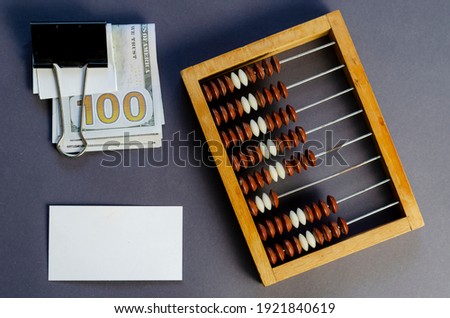 One Hundred Dollar Bill Sandwiched Binder Clip, Blank Business Card, and Old Wooden Abacus on Gray Background. Top view, flat lay.