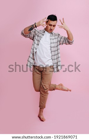 A man in casual clothes dances barefoot standing on one leg, pictured on a pink background. 
