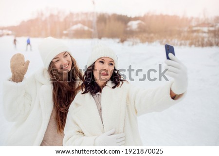 Two beautiful caucasian young woman with white hats taking selfie photo with smartphone outdoors in winter. Concept friendship lifestyle.