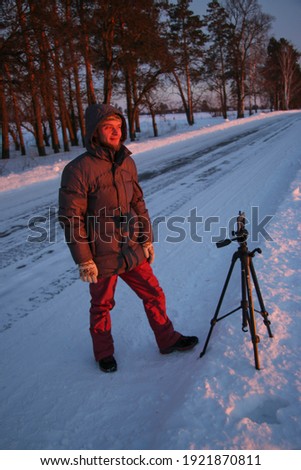 Man with phone on tripod making time-lapse photographs of winter zidio sunset. The field, road and trees are covered with snow.