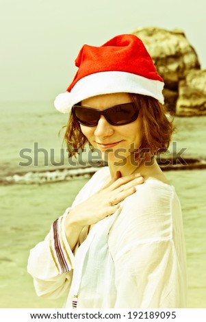Funny Santa Girl with sunglasses on tropical beach at rocks background. Beautiful brunette young woman smiling portrait in red Christmas hat celebrating New Year. Conceptual image with vintage filter
