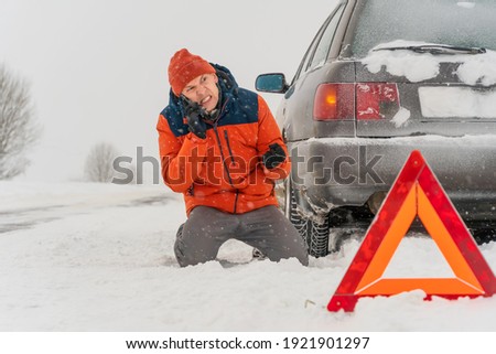 Angry young man in an orange jacket is on his knees talking on a mobile phone on a snowy road. He is in a state of stress because his car is broken. Winter travel, traffic problems, concept of care