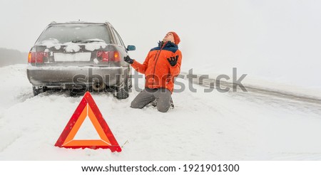 Angry young man in an orange jacket is kneeling on a snow-covered road. He is in a state of stress because his car is broken. Winter travel and recreation, road problems and assistance concepts.Banner