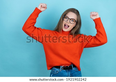 Pretty woman with long hair wearing a casual sweater and glasses over blue background very happy and excited making winner gesture with raised arms, smiling and screaming for success.