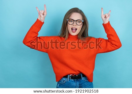 Pretty woman with long hair wearing a casual sweater and glasses over blue background shouting with crazy expression doing rock symbol with hands up