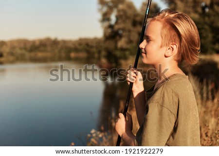 Young European blond boy fishing in summer sunny day, looking in distance, enjoying his spare time, wearing green t shirt, standing on bank of river near water. Royalty-Free Stock Photo #1921922279
