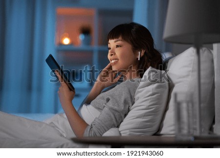 technology, internet and people concept - happy smiling young asian woman with tablet pc computer and wireless earphones lying in bed at home at night