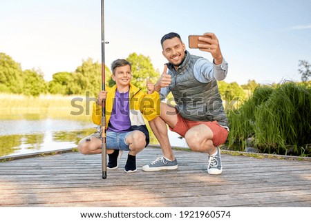 family, generation, summer holidays and people concept - happy smiling father and little son with fishing rods taking selfie with smartphone and showing thumbs up on river
