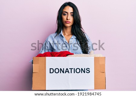 Beautiful hispanic woman volunteer holding donations box relaxed with serious expression on face. simple and natural looking at the camera. 