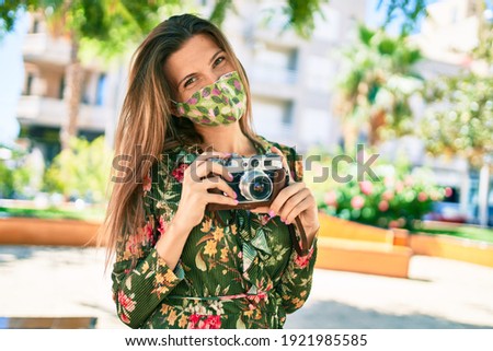 Beautiful caucasian woman outdoors on a sunny day wearing coronavirus safety mask taking pictures using vintage camera