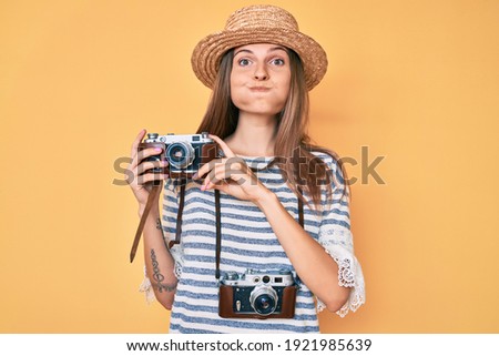 Beautiful caucasian tourist woman holding vintage camera puffing cheeks with funny face. mouth inflated with air, catching air. 