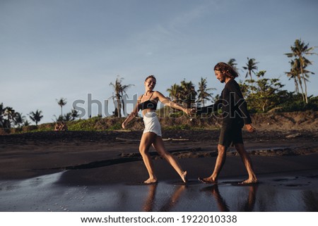 on the black coastal sand runs a girl and pulls the hand of a man walking in a step against the background of palm trees. High quality photo