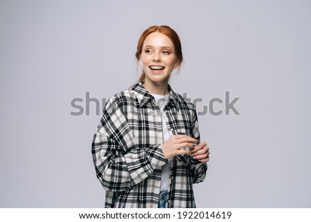 Cheerful young woman wearing wireless earphones listening popular song on isolated white background, looking away. Pretty redhead lady model emotionally showing facial expressions.