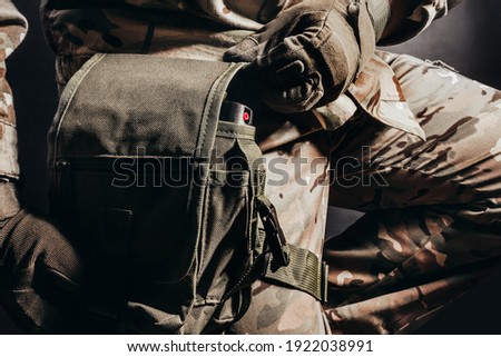 Photo of sitting soldier in camouflaged uniform and tactical gloves using leg bag on black background.