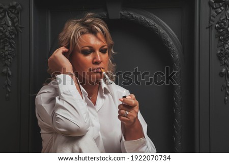 Portrait photo cute middle aged 45 years old blonde woman in domestic room. Female in white clothes in dark interior. Barefoot with cigarettes. Indoors positive confident mature woman. Copy space