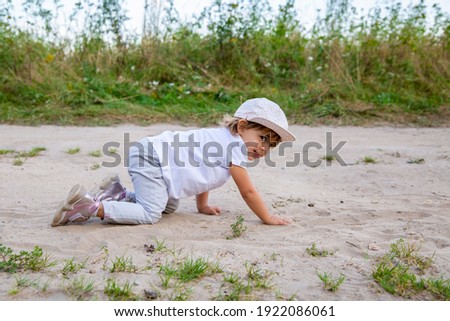 cute happy toddler in a cap crawls on all fours along a country road in the sand on a sunny summer day.