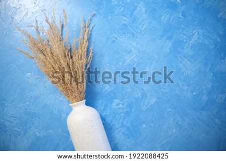 The composition consists of a white ceramic vase with a bouquet of dried spikelets, a golden photo frame with a dried flower on a blue background painted with oil paints. Stylish home decor.