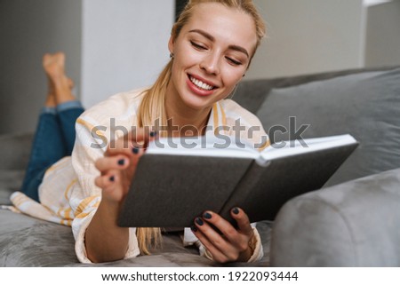 Happy woman smiling and reading book while lying on couch at home